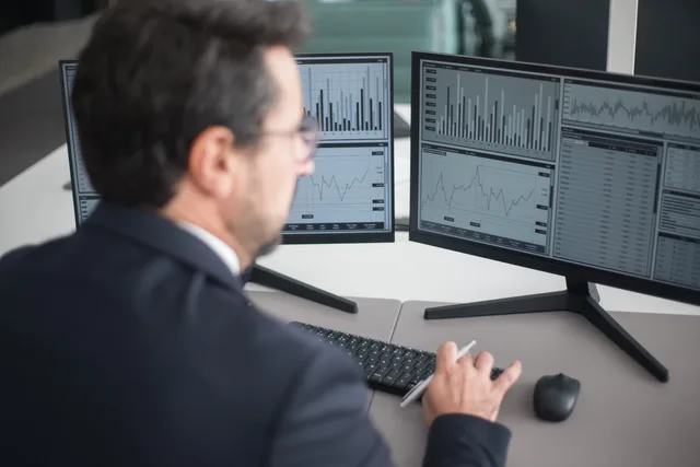 A businessman in a suit analyzing financial charts and graphs on two computer monitors indoors. Kvartalsdata i praktiken: så följer du svensk efterfrågan